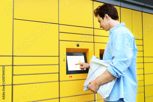 man uses a yellow parcel locker terminal for a contactless delivery pickup. This self-service logistics technology simplifies ecommerce online shopping and shipping.