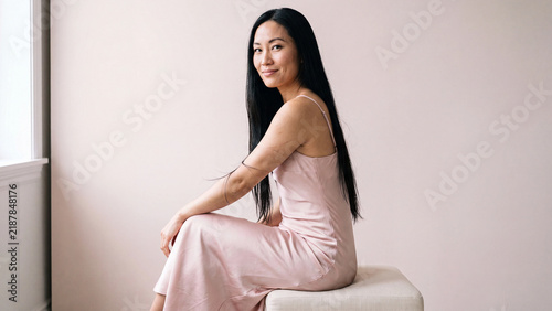 Brunette woman with long flowing hair wearing a pale pink satin dress resting her arm on a matching stool inside a softly lit contemporary room.