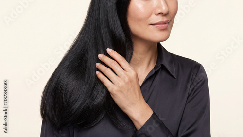 Woman touching her long black hair while wearing a dark satin blouse against a neutral studio background during a professional beauty photoshoot session.