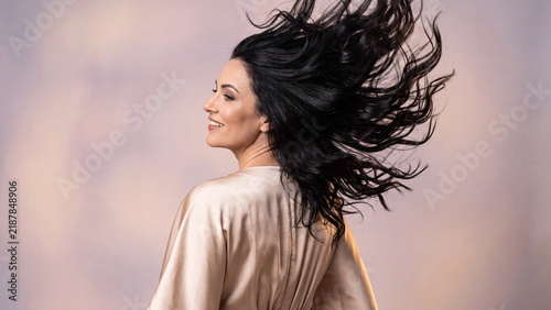 Brunette woman wearing a silky robe turns around with a joyful expression while her voluminous dark hair dynamically tosses in the air creating a dramatic motion effect.