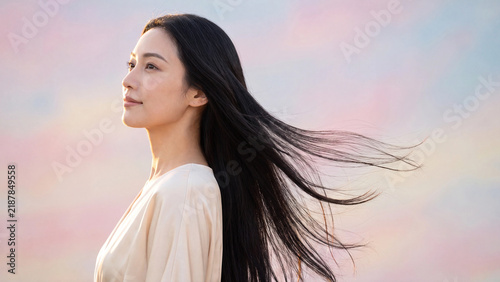 Woman with long dark flowing hair stands outdoors looking toward the horizon while the gentle breeze blows her strands gracefully against a pastel sky backdrop.
