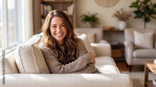 A happy woman with long wavy hair sits on a beige sofa in a bright living room and smiles at the camera with brown hair rests