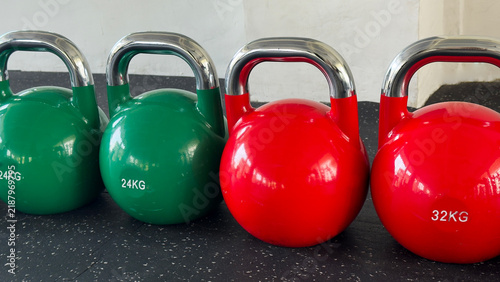 Green 24 kg and red 32 kg kettlebells with polished metal handles arranged in a row on a rubber floor, illustrating progressive strength, weight choice, functional fitness, and gym training.
