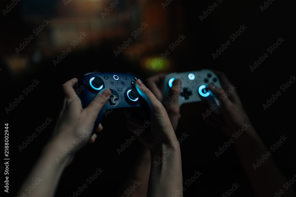 Fototapeta premium Close-up of couple hands holding illuminated wireless game controllers, actively competing in online video game tournament, enjoying leisure time and modern digital entertainment together at home.