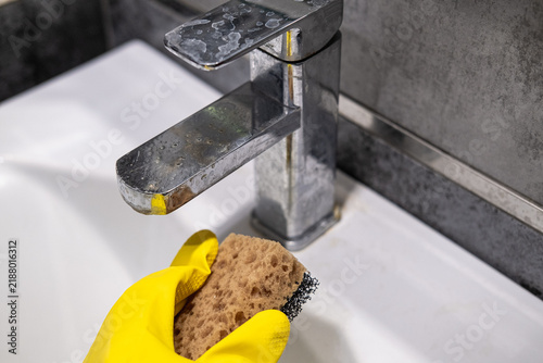 Cleaning the bathroom sink, she is polishing the faucet using a sponge, protective yellow gloves and spray detergent.