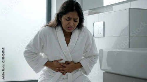 Young woman in bathrobe holding stomach in pain suffering from diarrhea and abdominal cramps in bathroom, medical illness and digestive discomfort concept video, medium shot selective focus