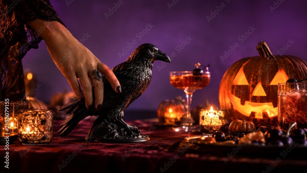 Obraz premium Female hand in black lace touching decorative raven on Halloween table. Glowing jack o lantern pumpkins, candles and cocktails on purple background.