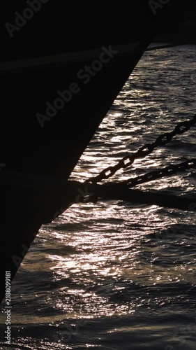 Silhouetted boat hull and chain with golden sunlight reflecting on calm, shimmering water. Serene and peaceful vertical footage.