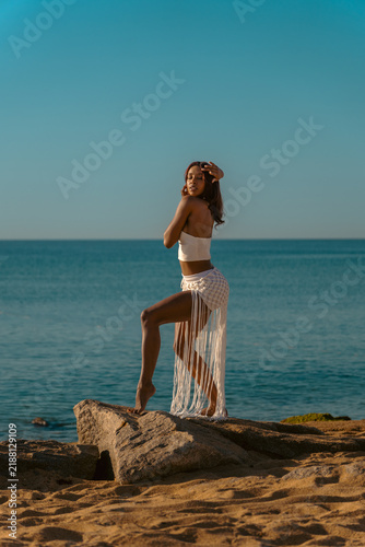 Konstfotografi A beautiful Cuban woman posing sensually in a suggestive dance on the beach rocks by the sea, wearing a white summer outfit