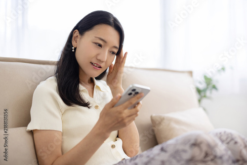 Young Japanese woman frowning and touching her head while looking at a smartphone on a sofa