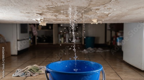Water leaking from damaged ceiling into a blue bucket on kitchen floor during plumbing emergency