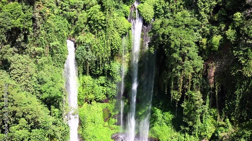 Waterfalls pour down forest cliff, framed by thick tropical greenery and bright moss. Waterfalls define waterfall landscape, with long white streams cutting through dense vegetation in daylight.