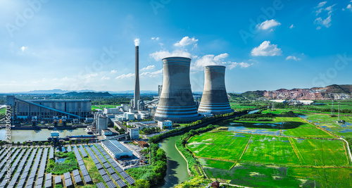 Aerial view of large cooling towers and solar panel arrays next to green fields under bright daylight and blue sky.