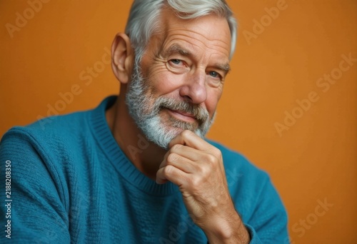 Older man with white beard, wearing sweater, smiling, in front of orange background.