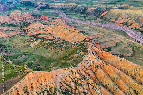 Canvastavla High-angle aerial view of rugged orange sandstone ridges, green grassy slopes, and a dry riverbed under soft natural daylight in Xinjiang
