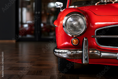 Close-up low angle shot of a classic red vintage sports car headlight and chrome bumper in a showroom, showcasing retro automotive design, elegance, and luxury collection.