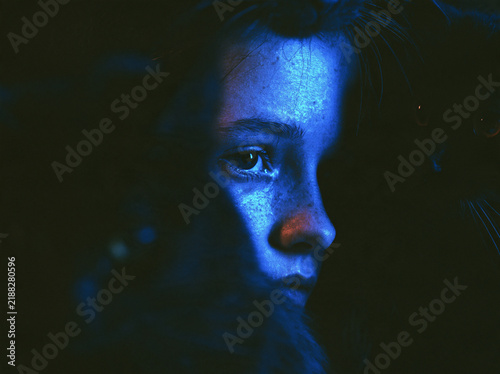 Close-up portrait of a young woman in the dark illuminated by striking blue light with a black cat beside her