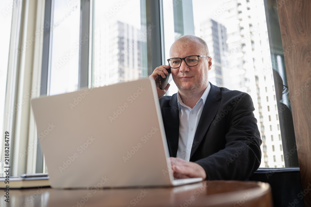 Obraz premium Mature businessman talks on smartphone while using laptop in modern office, representing multitasking, client communication and digital work.