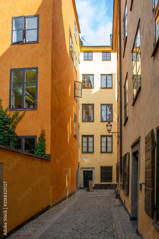 Fototapeta premium Narrow cobblestone alley of Skepper Olofs Gränd in Gamla stan, Stockholm. Historic yellow and orange buildings with bright sun glare reflection in windows under a blue summer sky.