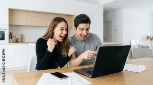 Excited young couple celebrating mortgage loan online approval success pointing at laptop screen in kitchen at home