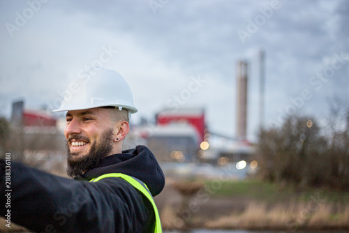 Male engineer smiling, wearing hard hat and safety vest, looking away from camera with an industrial power plant in background