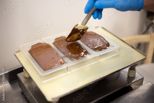 Handmade artisan chocolate bars being decorated with melted chocolate by a confectioner wearing protective gloves. Several chocolate pieces are arranged on a tray in a confectionery workshop, showing