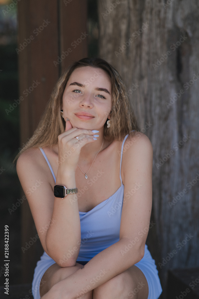 Fototapeta premium A beautiful young woman in a light blue dress with thin straps sits outdoors, resting her chin on her hand, and smiles gently.