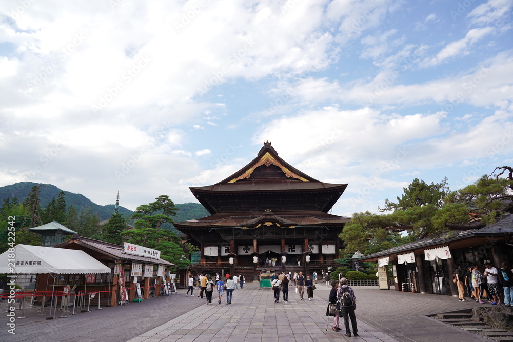 Obraz premium 日本の古都にある荘厳な寺院の境内が広がる夏の風景。中央には伝統的な木造建築の本堂が堂々と建ち、多くの参拝客や観光客が石畳の広場を散策している。背後には緑豊かな山々が連なり、穏やかな雲が浮かぶ青空が広がる長野県善光寺の一日。