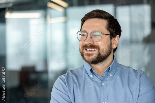 Smiling bearded man wearing glasses and blue shirt in modern office portrait with soft natural light, confident professional expression, blurred background and relaxed corporate atmosphere