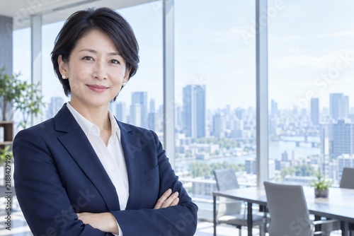Plakat 高層ビルを望む窓際の会議室で腕を組んで穏やかに微笑むスーツ姿の日本人女性