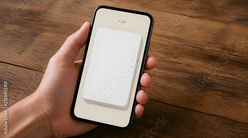 Hand holding smartphone with blank white mockup card on screen over wooden table surface for app interface presentation.