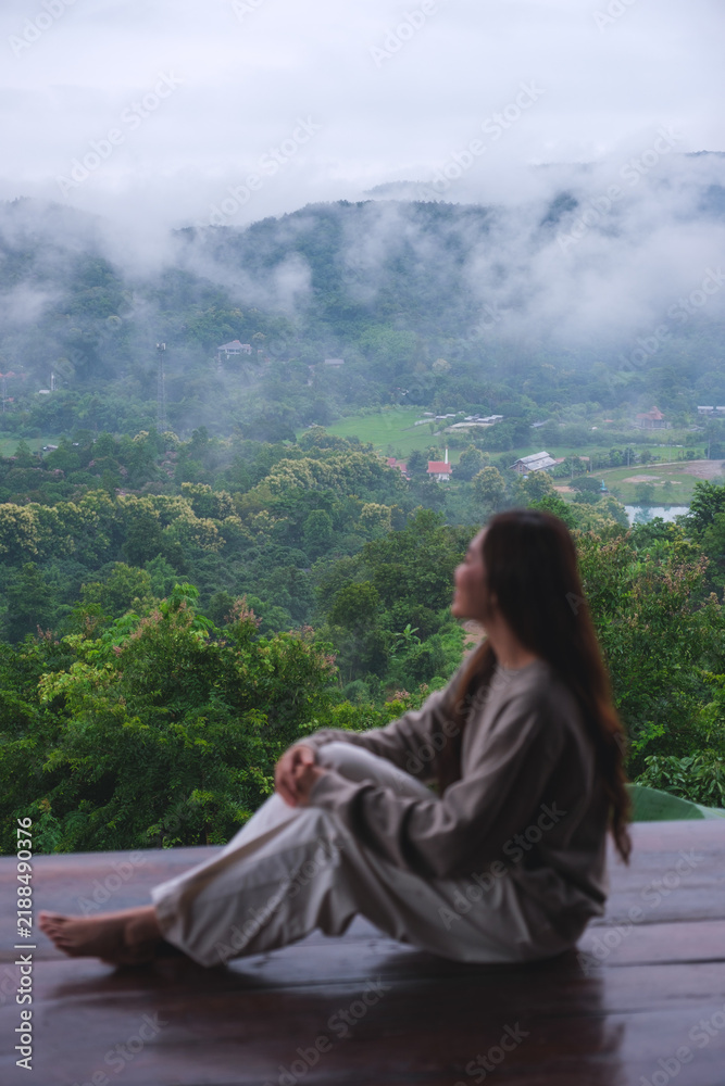 Obraz premium Vertical image of a woman sitting on wooden balcony with a beautiful mountain views on cloudy morning