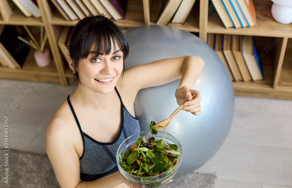 Fototapeta premium Woman eating salad celebrates healthy fitness nutrition. At home after training, she enjoys leafy greens by an exercise ball. Ideal for wellness content. Healthy lifestyle and diet concept.