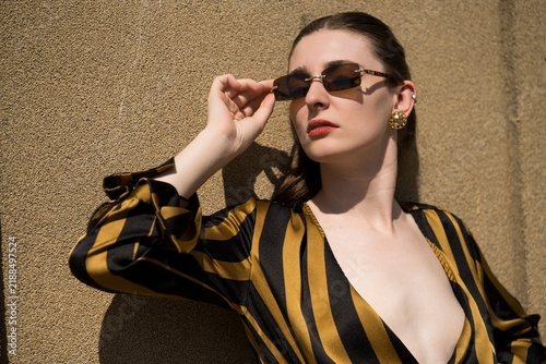 Portrait of attractive young caucasian woman wearing yellow and black striped blouse and sunglasses, standing near a wall on a warm, sunny summer day. Fashion, style, beauty.