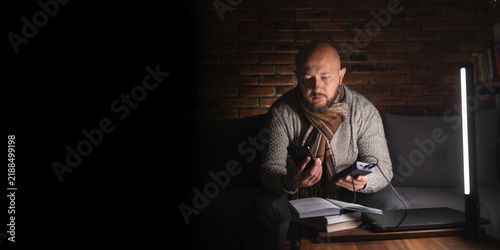 Ukrainian man on sofa in warm clothes using smartphone and holding power bank near led lamp, laptop and books on table during power outage in cold dark home caused by Russian attacks on energy grid