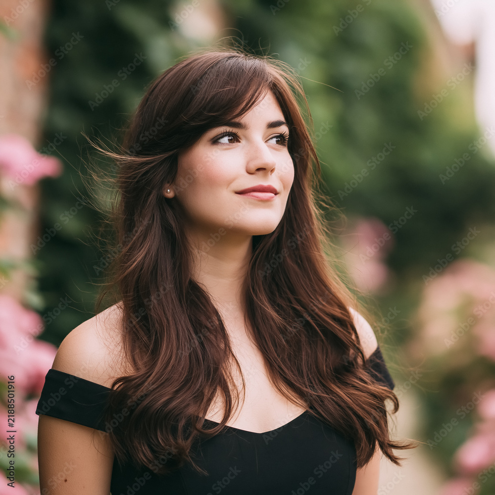 Fototapeta premium Young woman looking away, smiling in an elegant black dress, hair flowing in a garden with blooming pink flowers