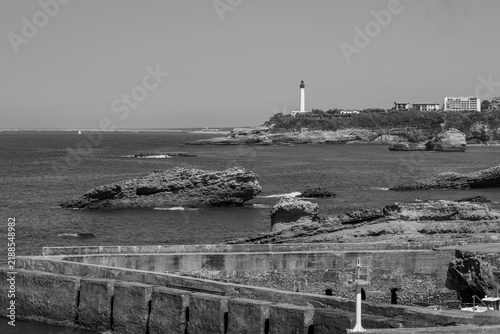 Black and white, A picturesque coastal scene in Biarritz France featuring rugged rock formations, a calm blue sea, a stone breakwater, and a distant lighthouse under a clear sky.
