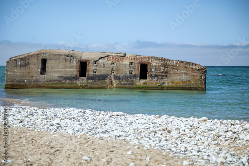 Fotografie An old concrete military bunker partially submerged in clear sea water at Vigso beach, showing a rusty entrance and weathered walls with green algae
