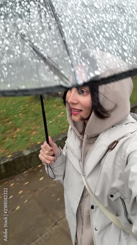 Side profile view of a cheerful young woman walking outdoors under a transparent umbrella during a rainy autumn day