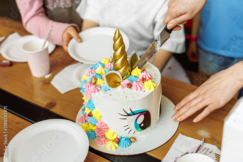 Cutting a colorful unicorn birthday cake with a golden horn for children. Festive sweet dessert concept at a kids party.