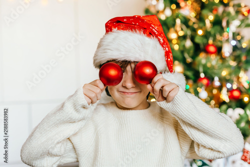 Funny boy in a Santa hat covering his eyes with red Christmas baubles. Playful holiday childhood concept at home.