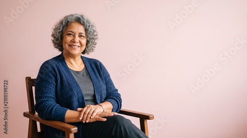 Smiling senior woman with curly gray hair sitting in a wooden chair against a pink background