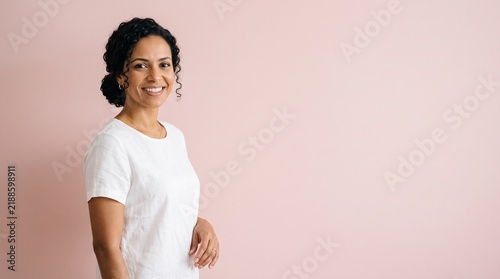 Smiling woman with curly dark hair and a white t-shirt against a soft pink background