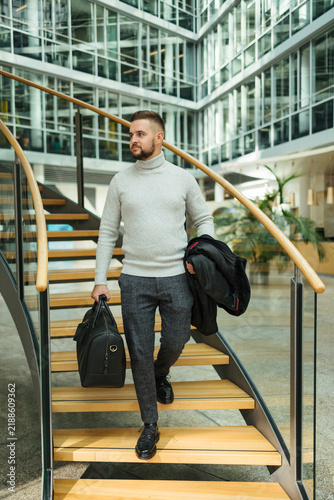 Portrait of handsome man in coat walking though the big office. Guy hold travel bag. Stylish guy after working day