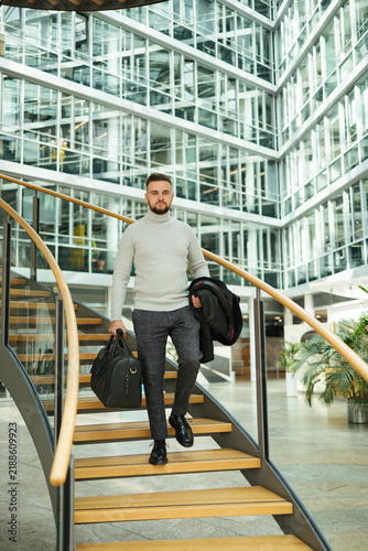Portrait of handsome man in coat walking though the big office. Guy hold travel bag. Stylish guy after working day