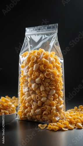 Delicious pasta in clear plastic bag, showcasing its vibrant yellow color and unique shape, surrounded by scattered pieces on dark background, evoking sense of culinary creativity