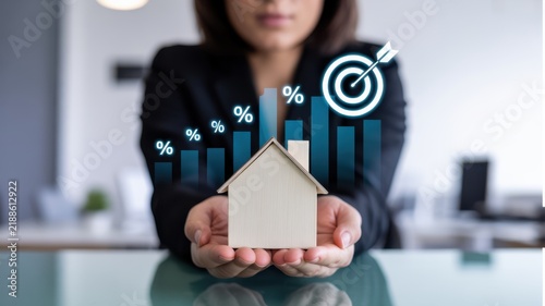 A Professional Holding A Model House With Rising Interest Rate Growth Charts