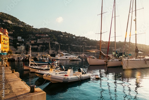 Sailboats and motorboats moored in calm Mediterranean waters along the waterfront in Menton, France. Warm sunlight illuminates the French Riviera coastline in September 2026.
