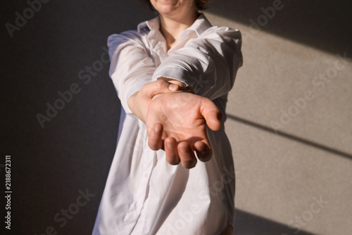Woman Stretching Arms Forward In White Shirt Against Sunlit Interior Wall With Strong Diagonal Shadows And Warm Late Afternoon Light.