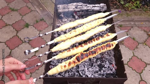 Close-up of hands turning skewers with sulguni cheese wrapped in golden puff pastry dough, baking on charcoal grill with smoke and flame